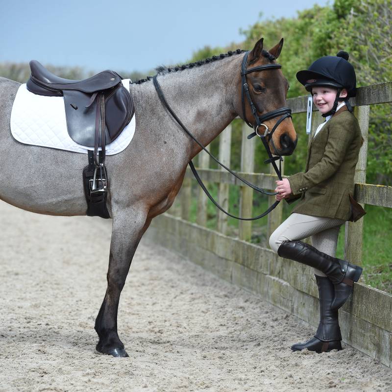 Smiling child in Dublin Albany Tweed Suede Collar Jacket stands by saddled horse outdoors.