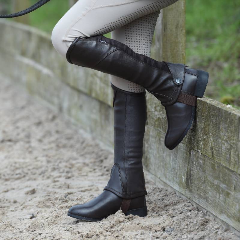 Person in brown boots, beige pants, and Dublin Stretch Fit Half Chaps stands by a wooden fence.