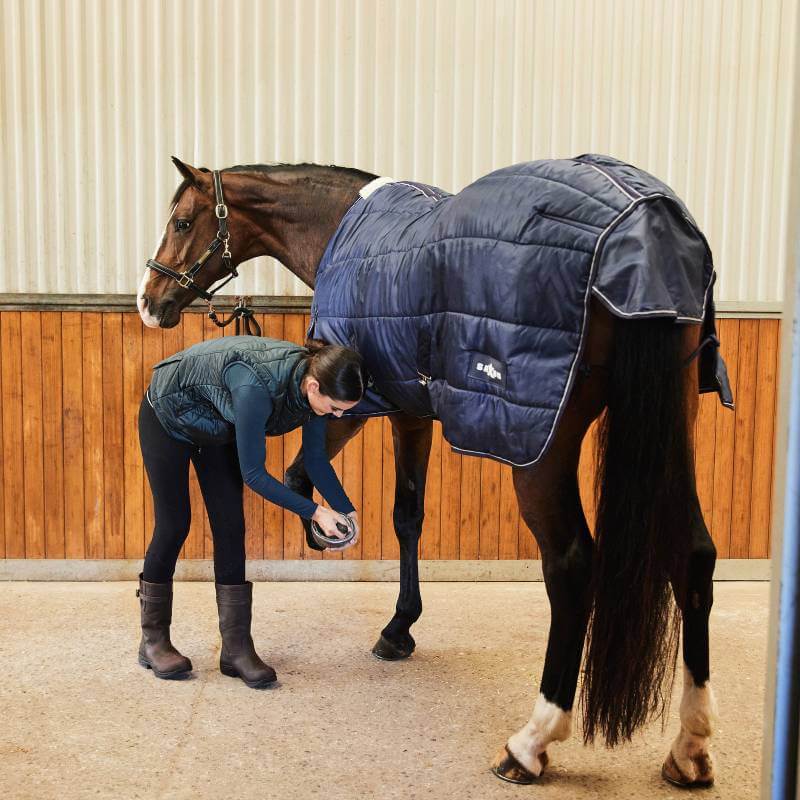 Person cleans a horse's hoof; horse in blue blanket, rider in Weatherbeeta Presley Puffer Vest.