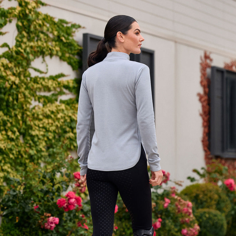 Woman wears Weatherbeeta London Layer Top Bluestone Marle outdoors by flowers and green plants.