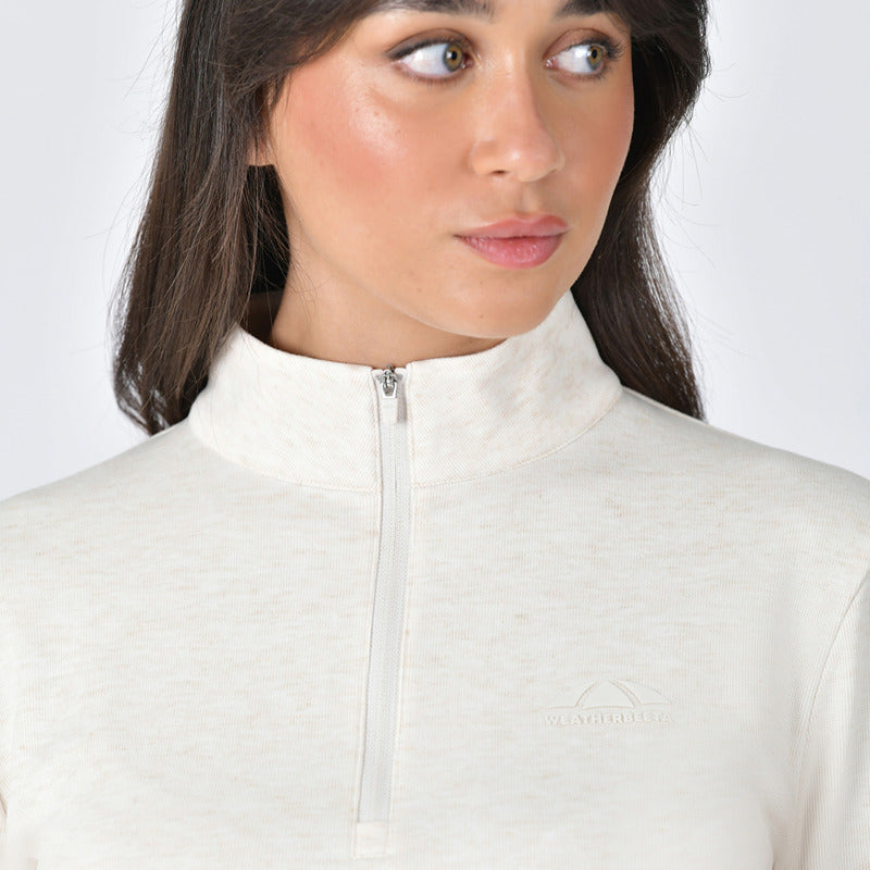 Woman in Weatherbeeta London Layer Long Sleeve Top Light Cream Marle, looking left, plain backdrop.