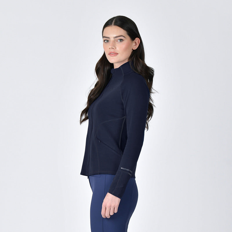 Woman in Weatherbeeta Copper Performance Jacket Navy and leggings stands sideways by plain backdrop.