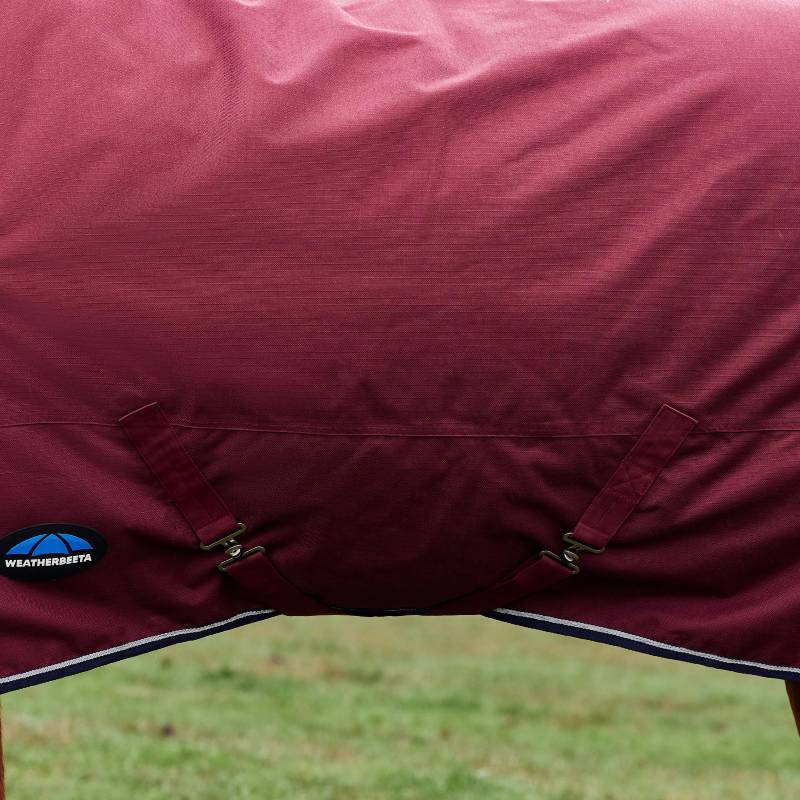 Close-up of a Weatherbeeta Comfitec 220g Burgundy/Navy turnout rug with straps on grass.