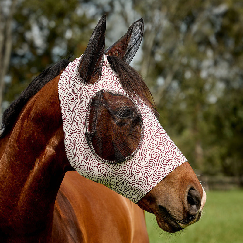 Brown horse in field wears Weatherbeeta Comfitec Deluxe Stretch Fly Mask; trees seen behind.