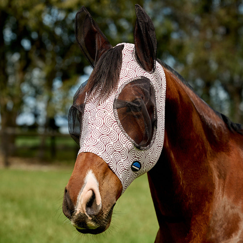 Brown horse in field wears Weatherbeeta Comfitec Deluxe Stretch Fly Mask; trees seen behind.
