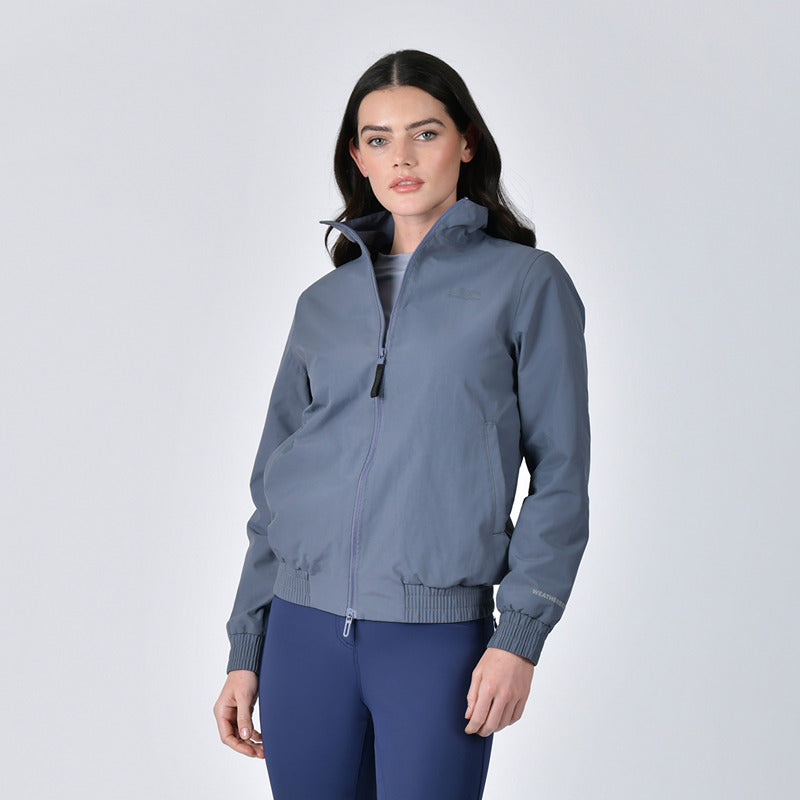 Woman in a Weatherbeeta Waterproof Team Jacket Slate Blue and navy pants against a light background.