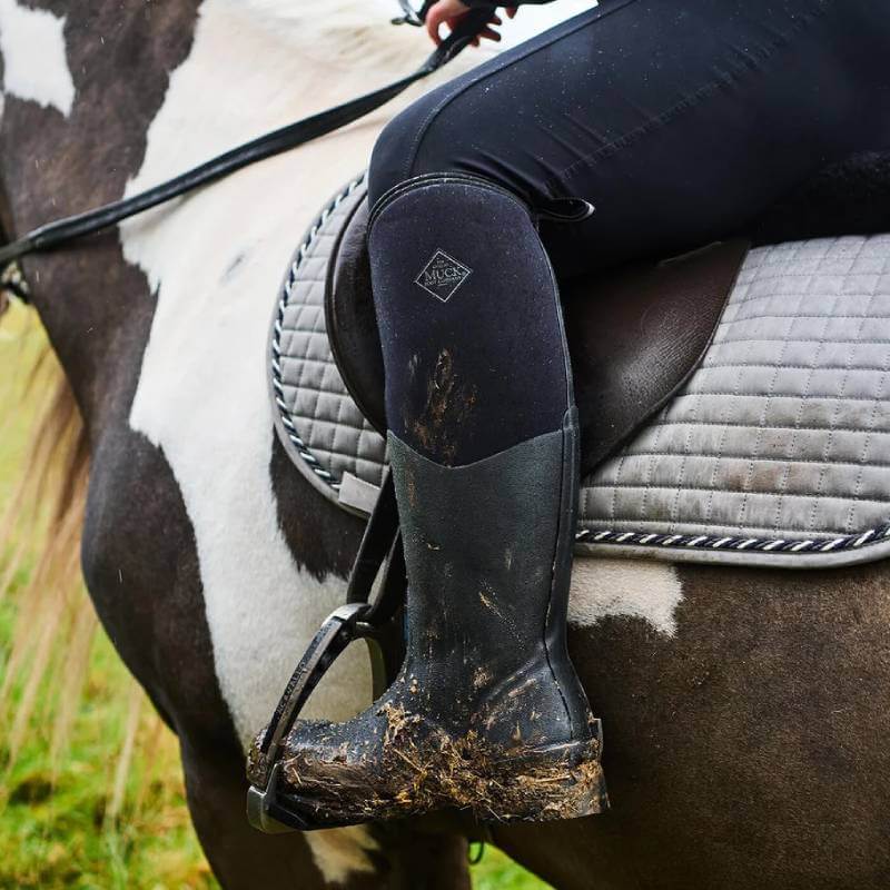 Close-up of a rider in Muckboot Colt Ryder Boots Black on a black and white horse with grey pad.