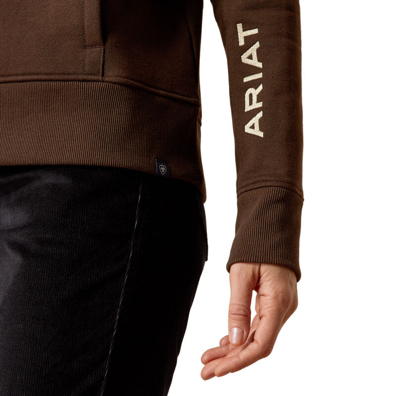 Person in a brown Ariat Womens Rabere Hoodie Mole with kangaroo pocket and ARIAT on the sleeve.