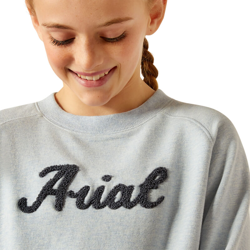 Girl smiling and looking down, wearing the Ariat Youth Benicia Sweatshirt in Cerulean Heather.
