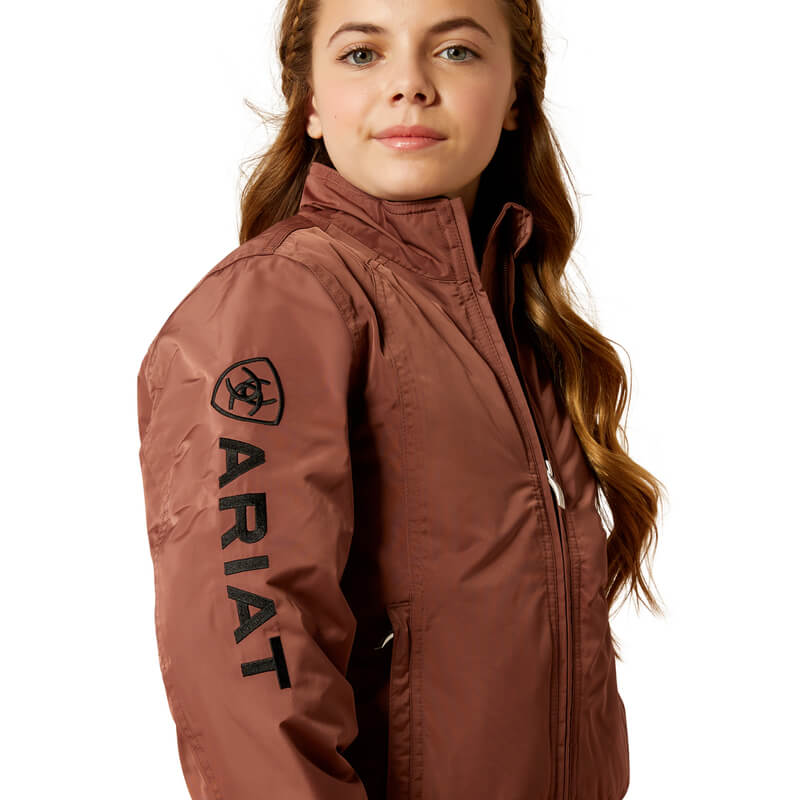 Girl in an Ariat Youth Stable Jacket Marron and jeans, smiling against a white background.