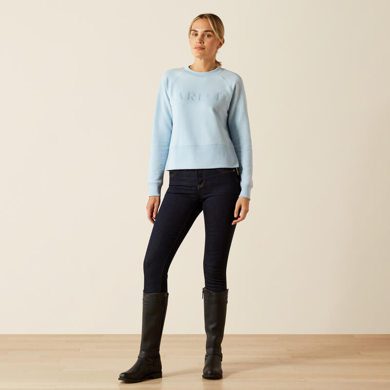 Woman wearing Ariat Womens Martine Sweatshirt Skyway, dark jeans, and black boots in a minimal room.