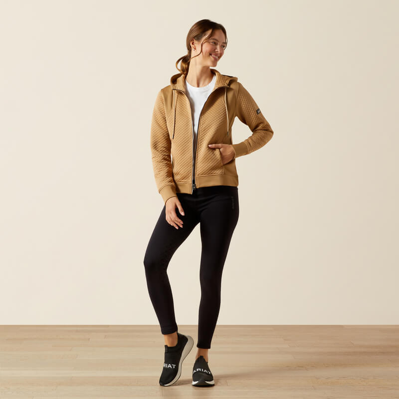 Woman in Ariat Womens Radiance Full Zip Tiger's Eye jacket, white shirt, and black leggings indoors.