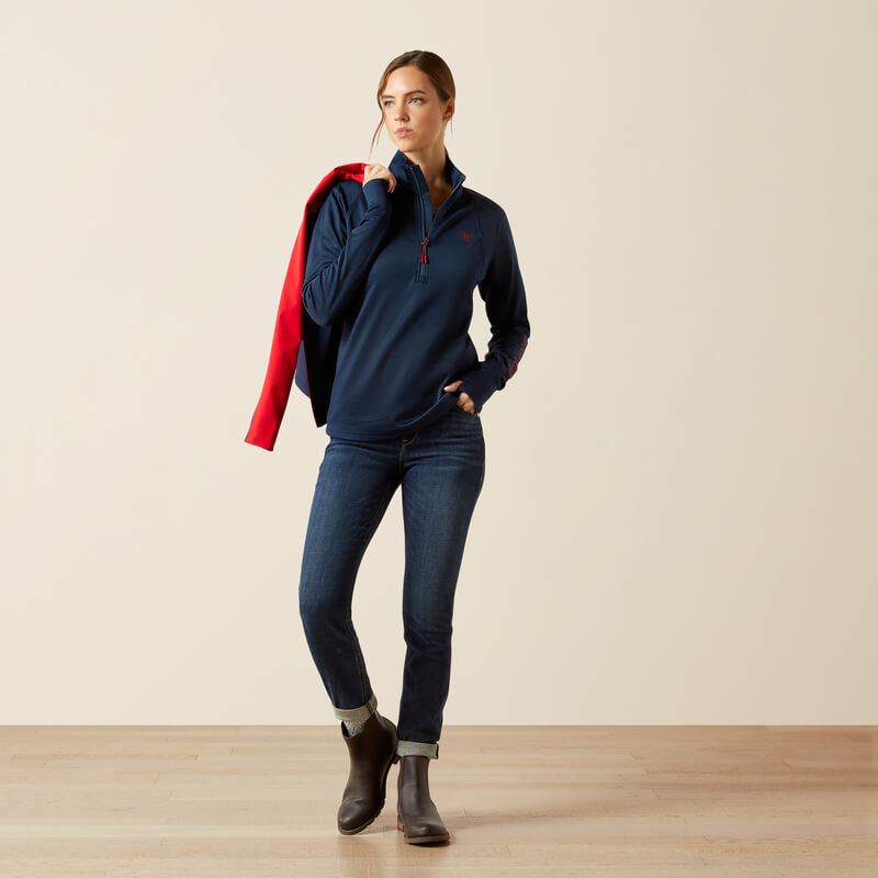 Woman in Ariat Womens Team EQ 1/2 Zip Pullover Navy holds a red jacket indoors.