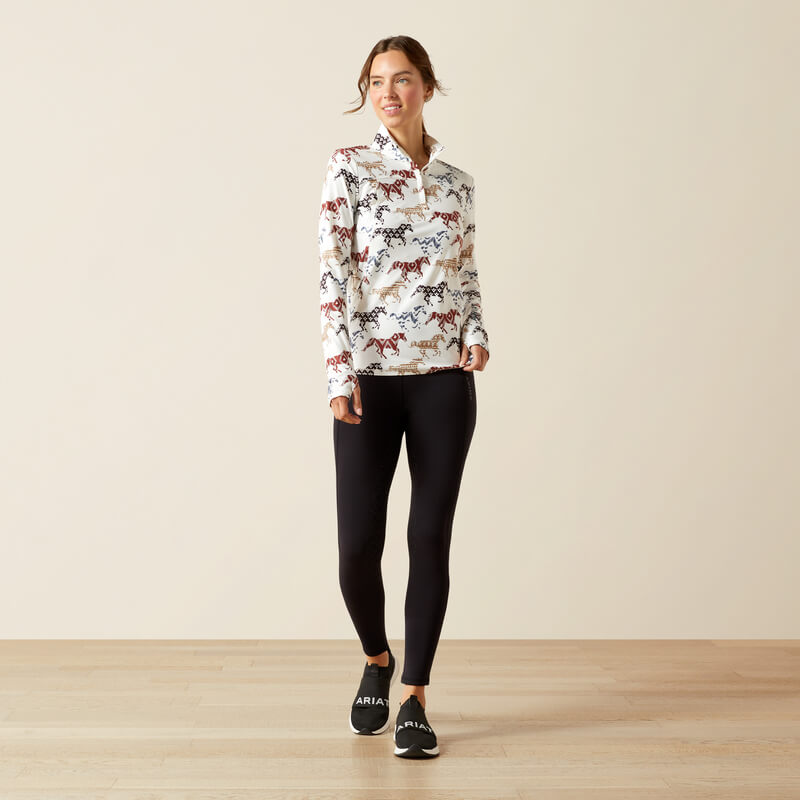 Woman in Ariat Lowell 3.0 1/4 Zip Painted Ponies baselayer, black leggings, and sneakers indoors.