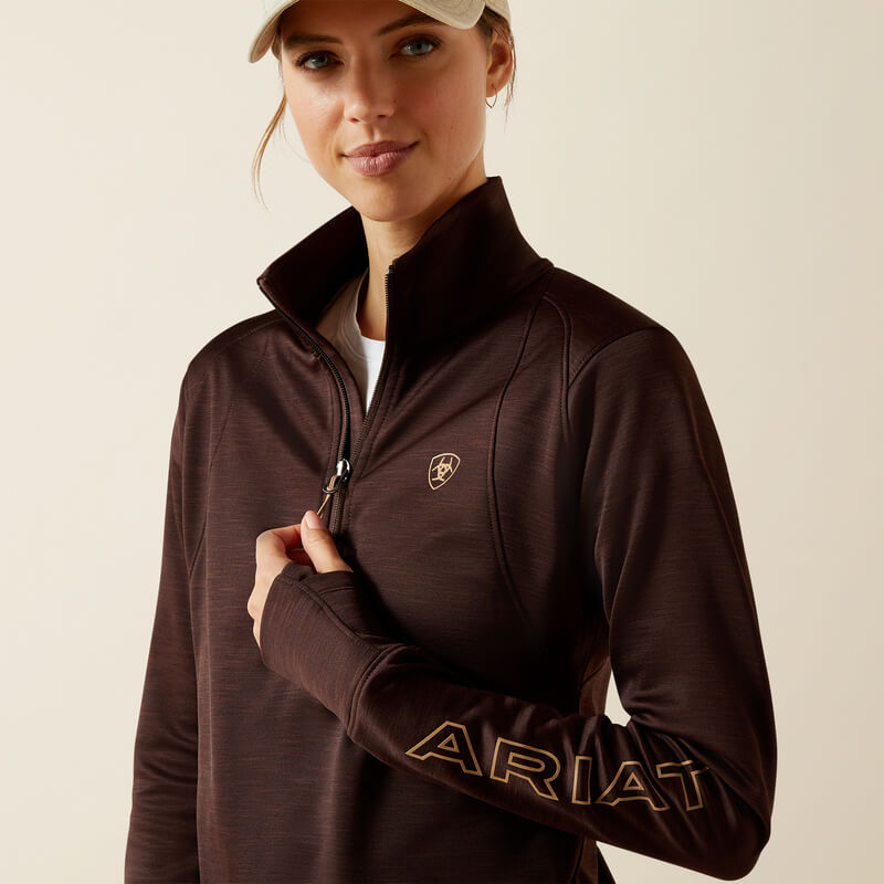 Woman in Ariat Womens Team EQ 1/2 Zip Pullover Java, jeans, and beige cap against a plain background.