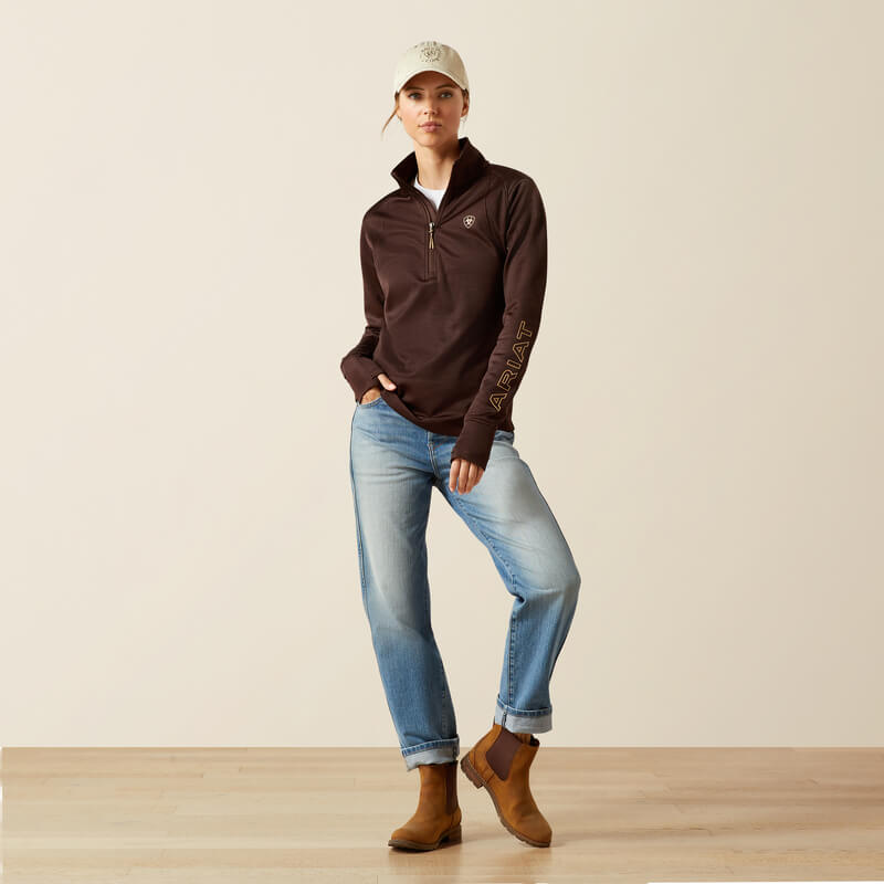 A woman in jeans, boots, and an Ariat Womens Team EQ 1/2 Zip Pullover Java stands against a plain background.