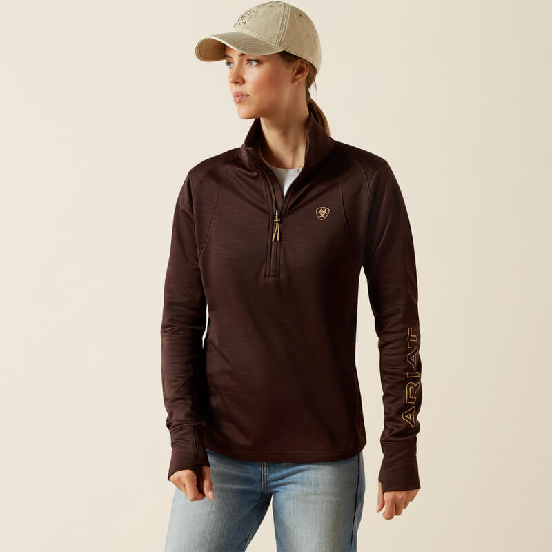 Woman in Ariat Womens Team EQ 1/2 Zip Pullover Java, jeans, and beige cap against a plain background.