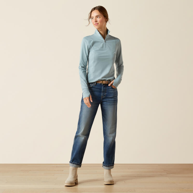 Woman in Ariat Gridwork 2.0 1/4 Zip Baselayer, jeans, and boots on wooden floor.
