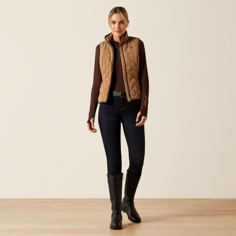 Woman in Ariat Womens Ashley 2.0 Insulated Vest Tiger's Eye, dark top, jeans, and black boots indoors.