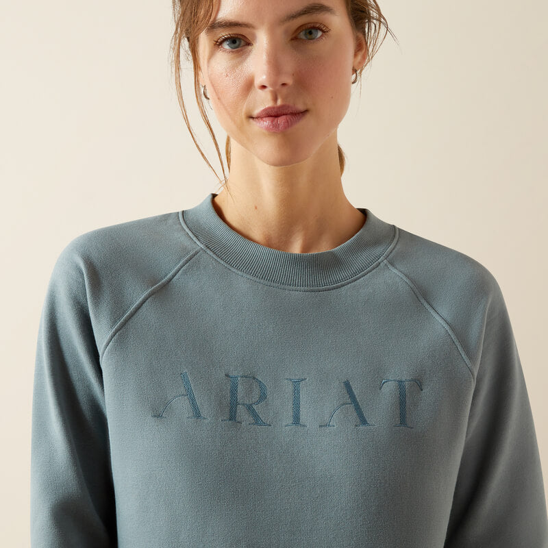 Smiling woman in an Ariat Womens Martine Sweatshirt Steel Sage and black pants stands against beige.