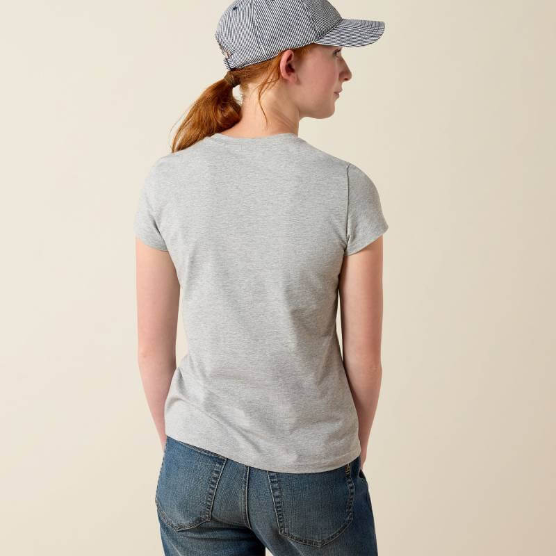 Person in Ariat Youth Vertical Logo SS T-Shirt, jeans, and cap stands with back to camera.