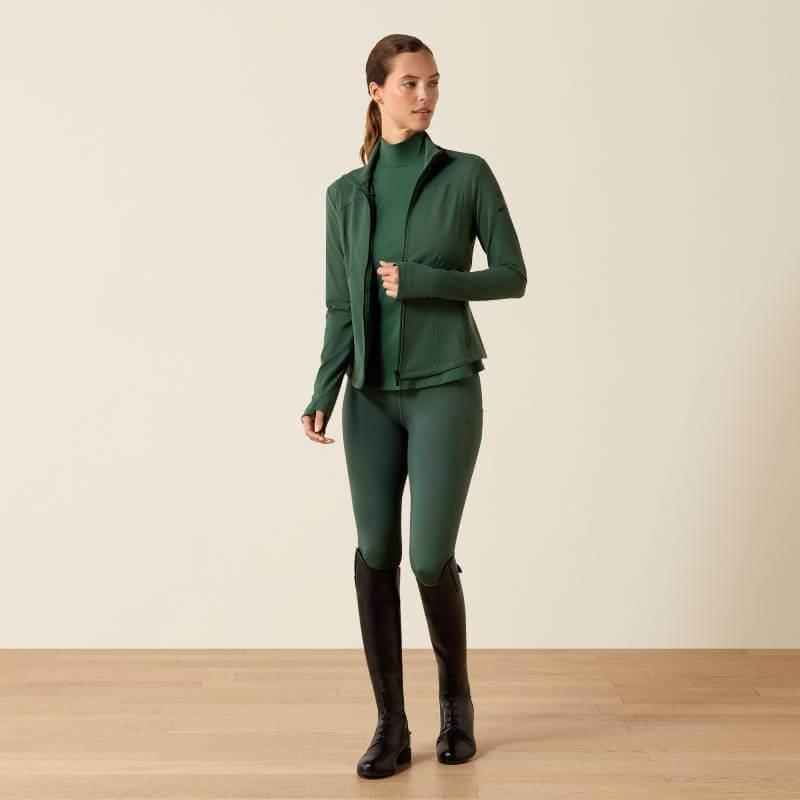 Woman in green Ariat Womens Solveil Full Zip Monstera and black boots stands on wooden floor.