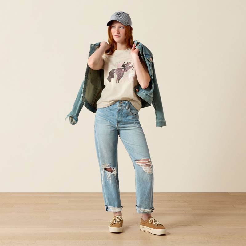 Person in jeans, Ariat Youth Floral Leap SS T-Shirt Oatmeal, denim jacket, cap, sneakers indoors.