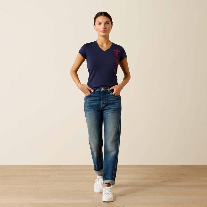 Woman in Ariat Womens Vertical Logo SS T-Shirt Navy/Red and jeans stands on wood floor.