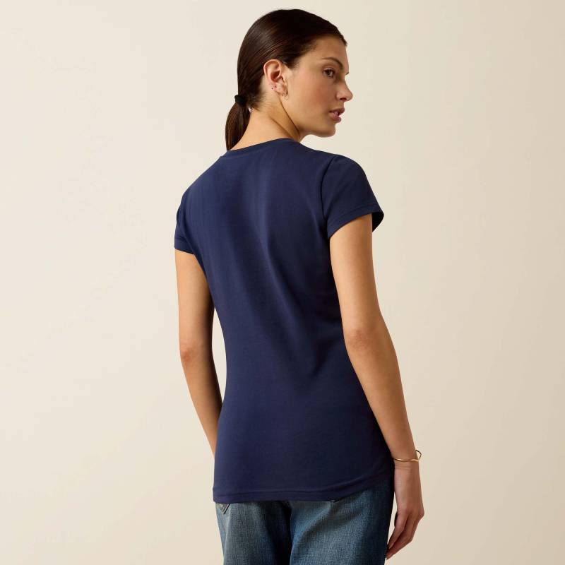 Woman in Ariat Womens Vertical Logo SS T-Shirt Navy/Red stands with her back, hair in ponytail.