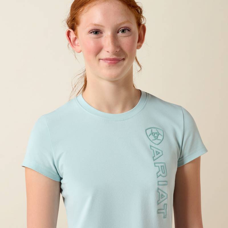 Girl in Ariat Youth Vertical Logo T-Shirt Starlight Blue/Blue Surf stands smiling, hand on hip.