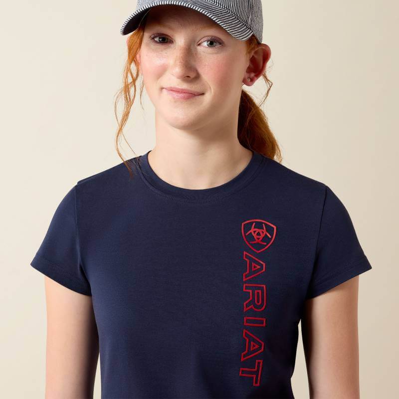 Teen girl in Ariat Youth Vertical Logo SS T-Shirt Navy/Red, hand in pocket, smiling brightly.