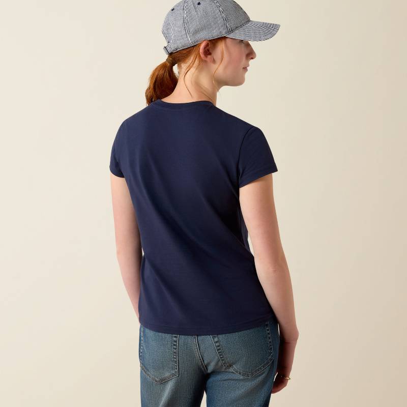 Person in Ariat Youth Vertical Logo SS T-Shirt Navy/Red, jeans, and cap stands with back facing.