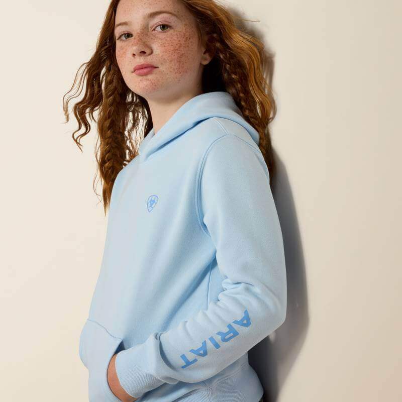 Smiling girl in Ariat Girls 2.0 Hoodie Nantucket Breeze, hands in pockets, beige background.