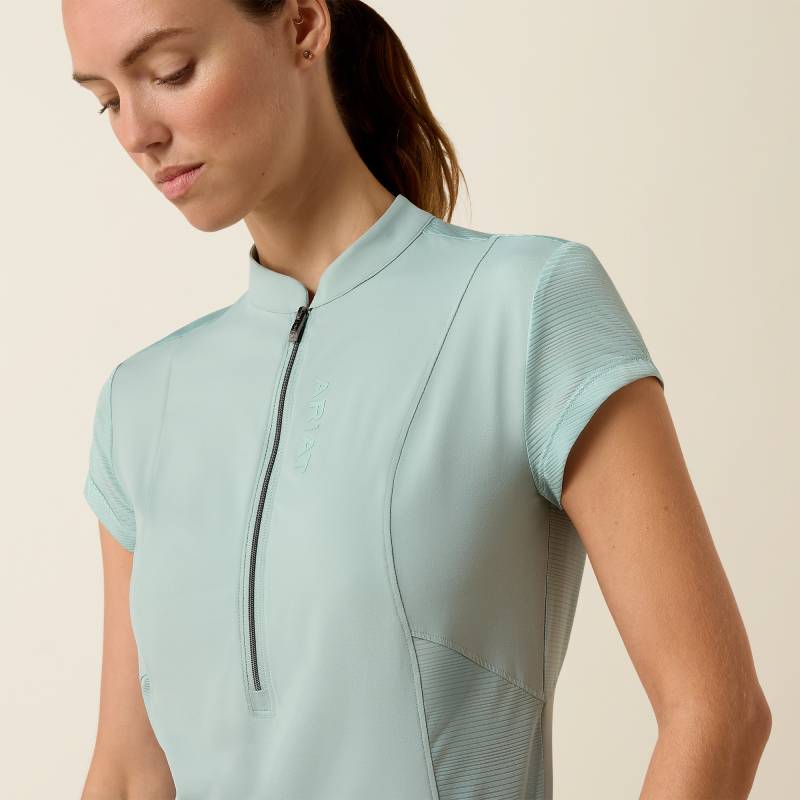 Woman in Ariat Breathe 1/2 Zip Cap Sleeve Baselayer Blue Surf, dark leggings, beige backdrop.