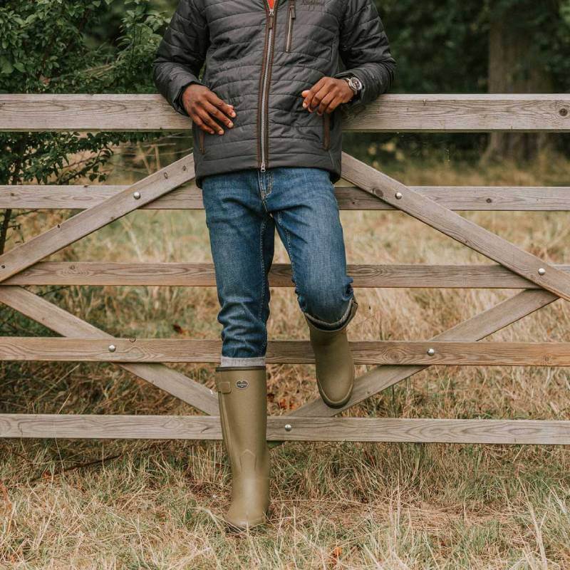 Man in jeans and Le Chameau Chasseur Neo boots leans on a wooden gate outdoors.