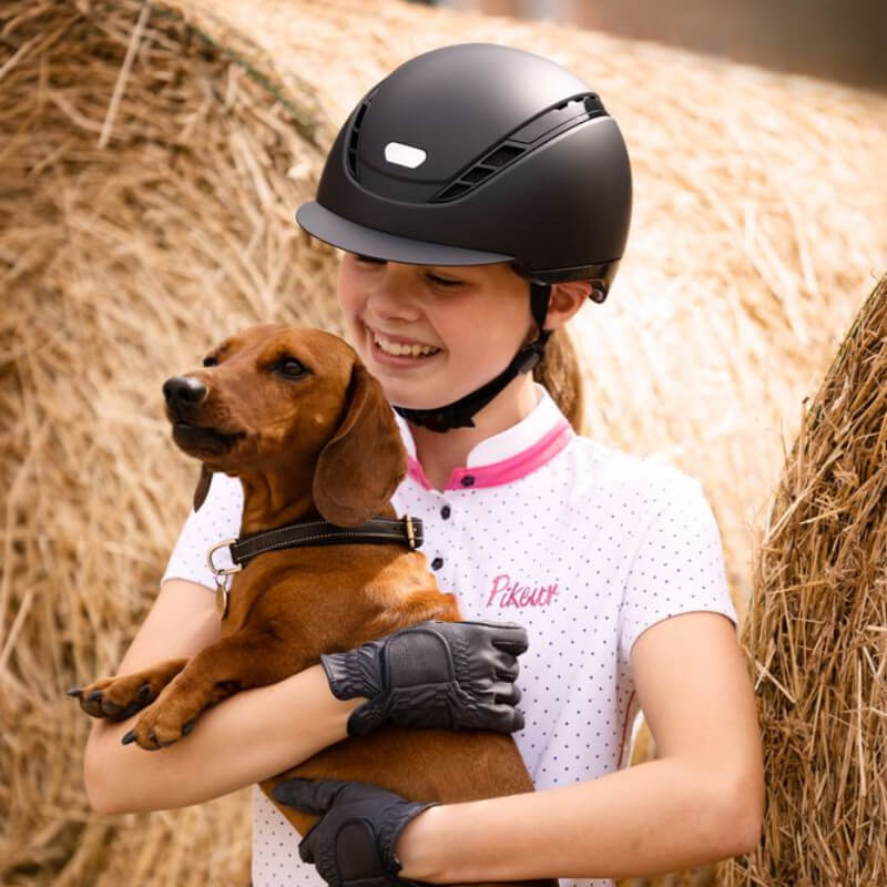 Girl in Pikeur Abus AirDuo Equestrian Riding Helmet holds brown dachshund before hay bales.
