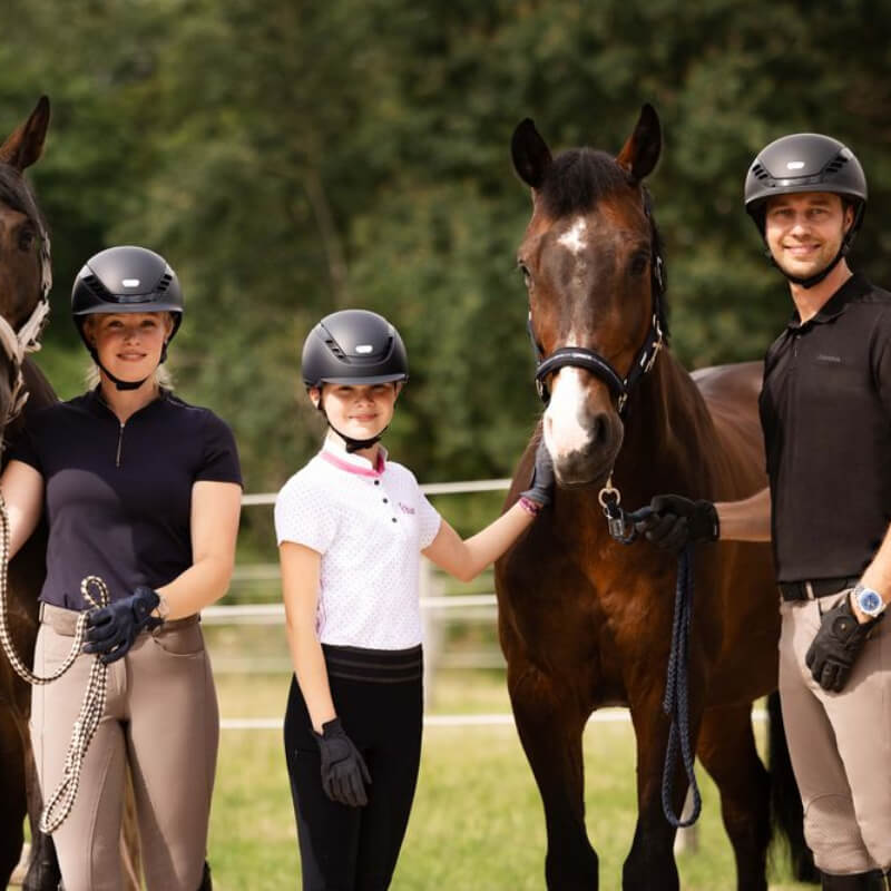 Three people in Pikeur Abus AirDuo Riding Helmets pose with two horses outdoors on a sunny day.