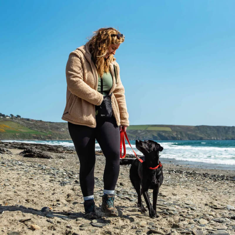 A woman with a Mountain Paws Dog Snack Pack walks a black dog on a rocky beach with ocean views.