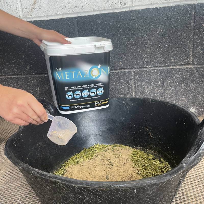 A person scoops NAF Five Star Metazone Powder 2.4kg into a black tub with feed.