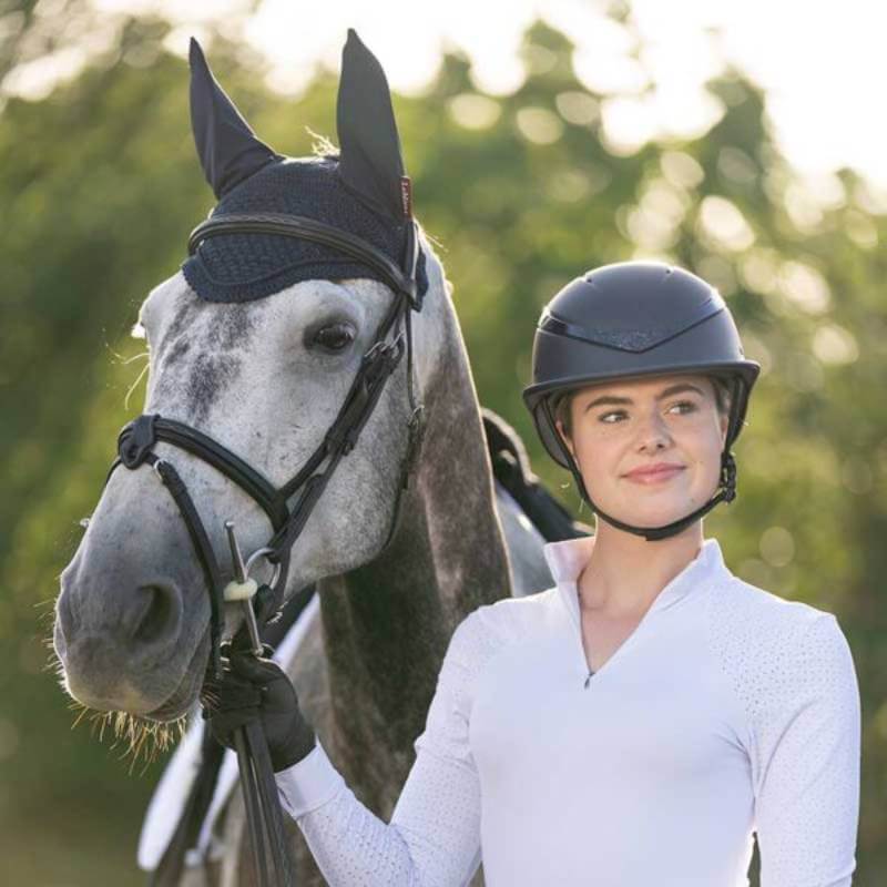 Smiling woman in riding gear wears Charles Owen This Esme Luna Midnight Blue by a gray horse.