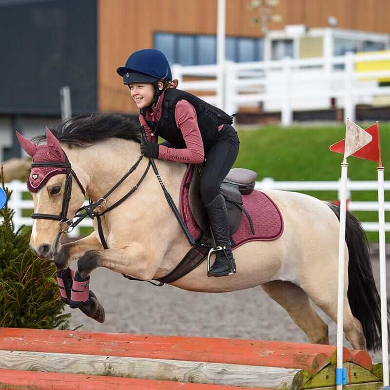 A child in a Charles Owen Kontor Black rides a horse over a red jump in an arena.