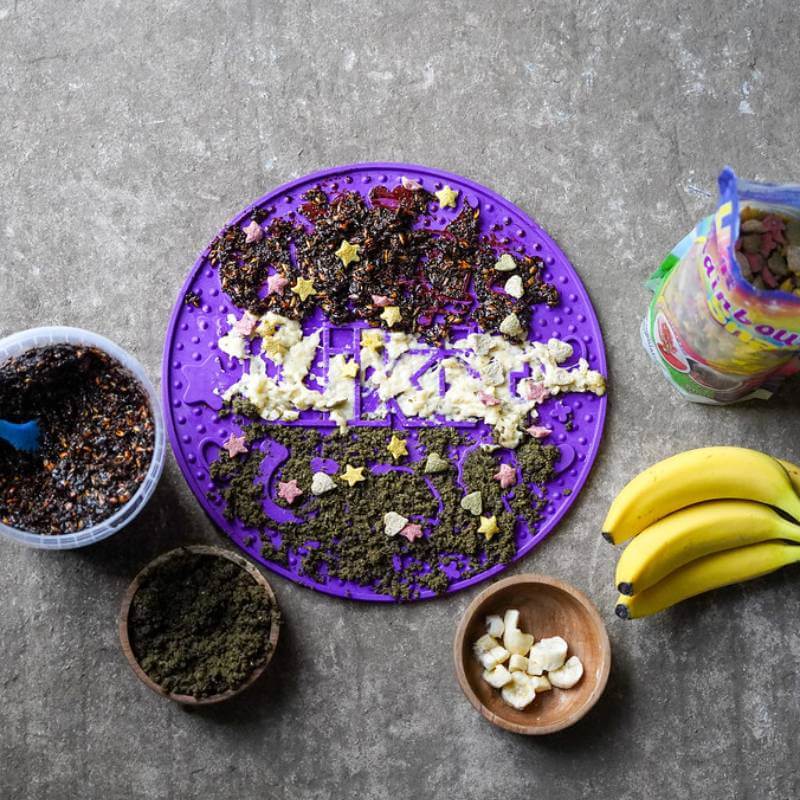 A Likit Graze Maze Purple slow feeder with treats, bananas & bowls on a gray surface.