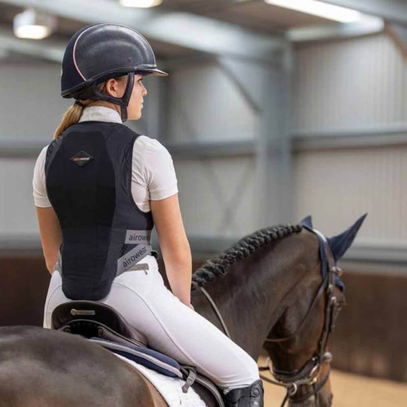 A rider dressed in white, seen from behind, wears a helmet and an Airowear Shadow Adults Black EN1621-2 certified back protector while riding a horse with a braided mane inside an indoor arena.