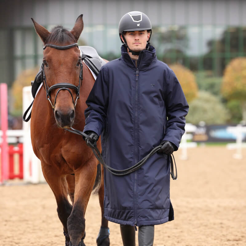 A person in an Aubrion Core All Weather Robe Unisex Navy leads a brown horse outdoors.