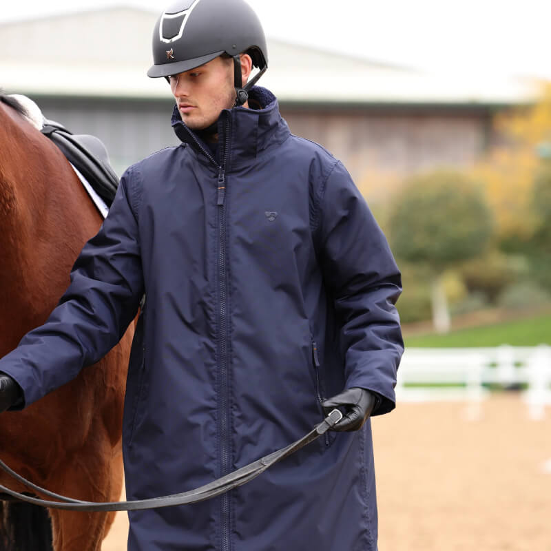 Person in Aubrion Core All Weather Robe Unisex Navy stands by a brown horse at an outdoor arena.