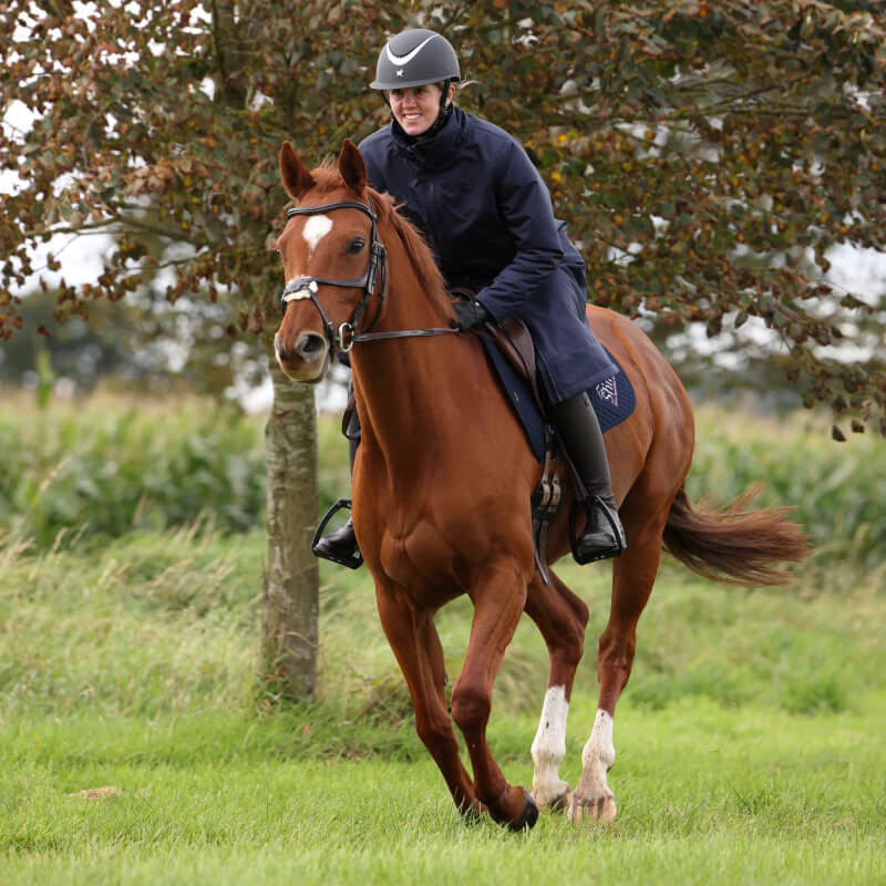 A person wears the Aubrion Core All Weather Robe Unisex Navy while riding a horse near trees.