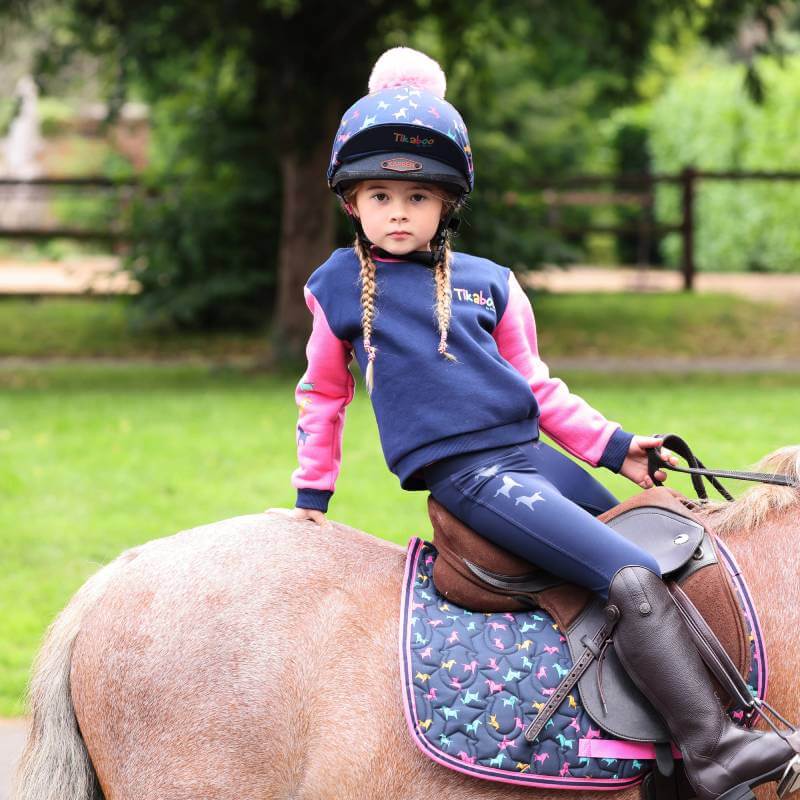 Young girl in Tikaboo Riding Tights Navy by Shires sits on a pony outdoors, wearing a helmet.