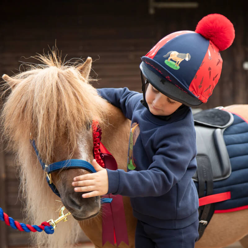 Child in Shires Winnie & Me Childs Hat Silk Navy/Red pets a brown pony with blue halter.