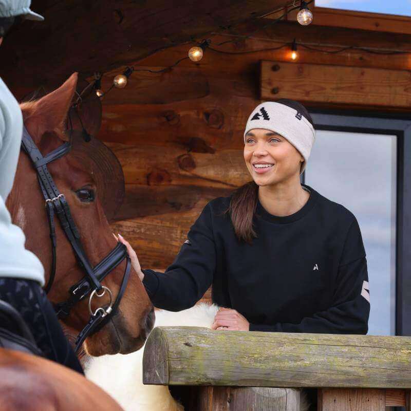 Smiling woman in Shires Aubrion React Sweatshirt pets a brown horse by a lit wooden building.