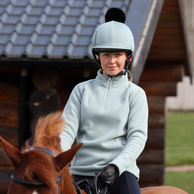 Wearing an Aubrion React Hat Cover Sage, a rider shows equestrian style on a brown horse outdoors.